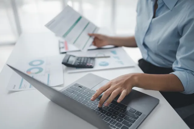 Person typing on a laptop beside printed charts and a calculator at a bright office desk