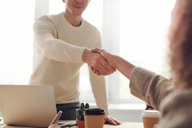 Two people shake hands across a table in a bright office with a laptop and coffee cups.